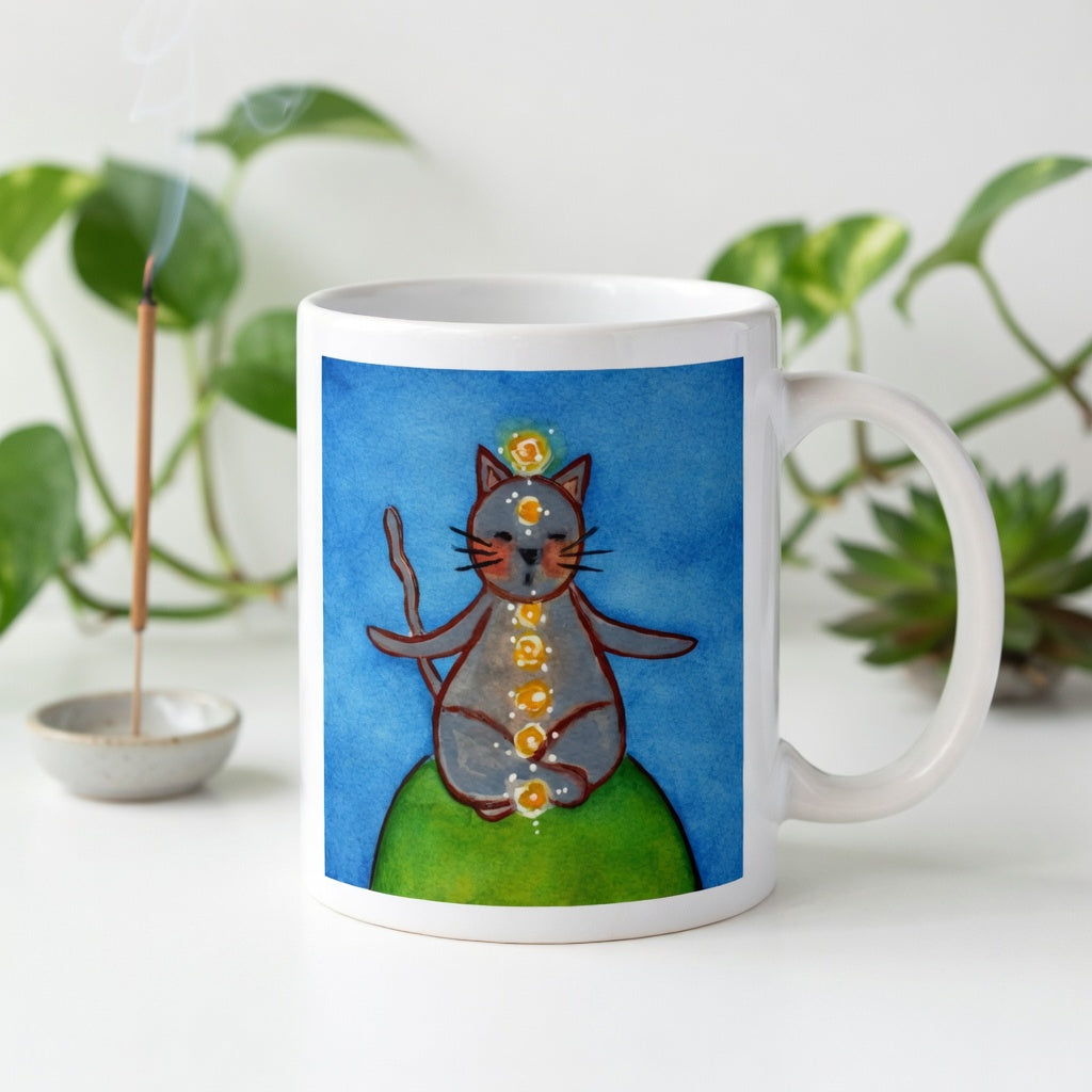 Gray cat meditating with glowing chakras on a deep blue calming background. Incense is burning behind the mug. There is a green potted plant in the background.