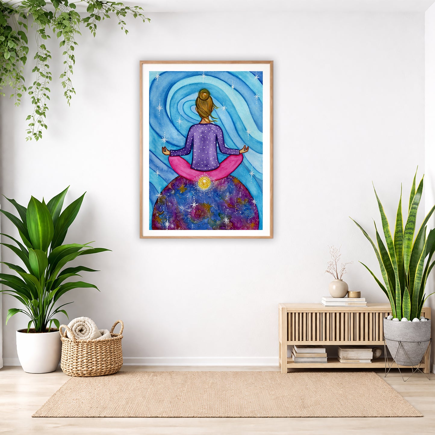 Poster of a woman meditating with her root chakra glowing. The poster is in the center of the living room wall. Below the poster to the left is a basket of material and a green floor plant. There is also a green hanging plant. To the right is a small table with books, a candle and a vase with dry flora. There is also a large green floor plant.
