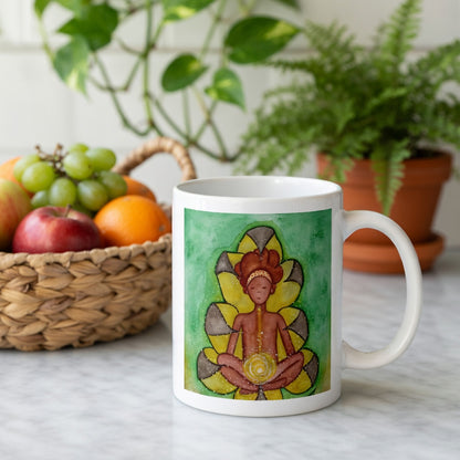Meditating figure with glowing sacral chakra below the navel, representing creativity, passion and emotional flow, spiritual chakra artwork on a coffee mug.  Behind the mug is a basket of fruit and a green potted plant.