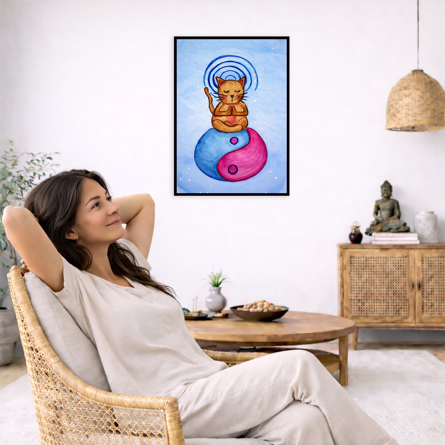Brown cat meditating on a yin yang symbol with its crown chakra glowing framed poster.  This is a print of the original hand-painted watercolor by Brazilian artist Thaís Suizo. The poster is in the center of the living room wall. To the left a woman is relaxing in her chair with her hands behind her head. There is a small coffee table beside her with a bowl of walnuts, incense and a small green plant. On the right side of the room is a cabinet with a Buddha statue, books and an urn on it. To the left is a g