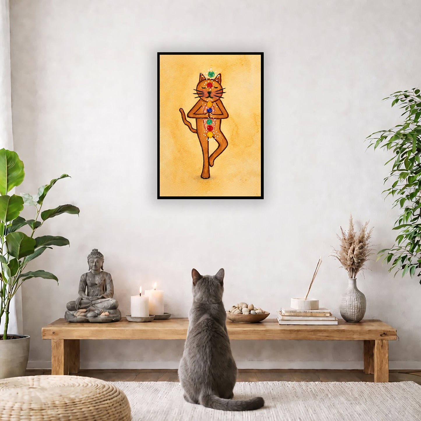 A brown cat standing in meditation with its chakras glowing framed poster.   This is a print of the original hand-painted watercolor by Brazilian artist Thaís Suizo.  The poster is in the center of the living room wall. A gray cat is facing the poster. In front of it is a coffee table with a Buddha statue and burning candle on the left and incense and an orange cat staring at the poster on the right. There is a green potted plant on both the left and right side of the coffee table.