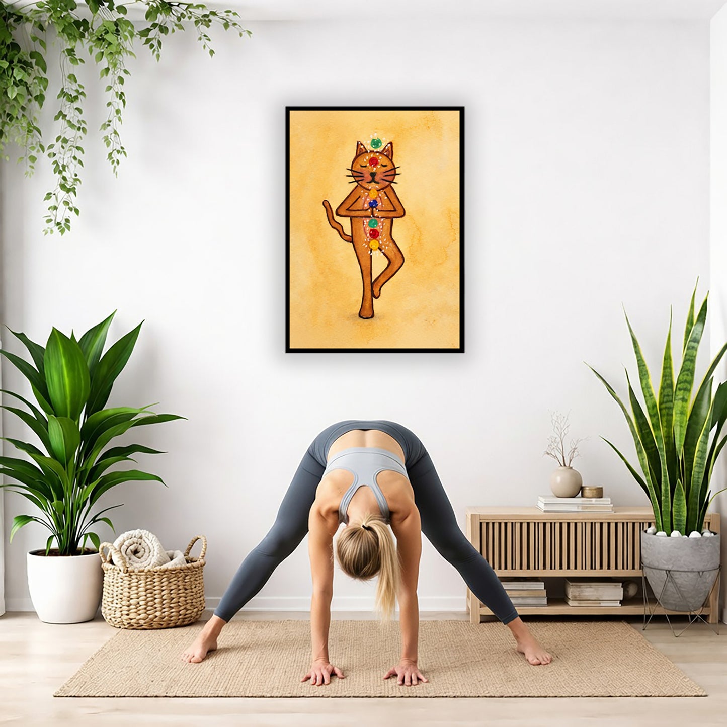 A brown cat standing in meditation with its chakras glowing framed poster.   This is a print of the original hand-painted watercolor by Brazilian artist Thaís Suizo.  The poster is in the center of the living room wall. A woman is doing a forward bend on her yoga mat. There is a padded bench below the poster with some books and a small potted plant on it. To the right and left are potted plants.

