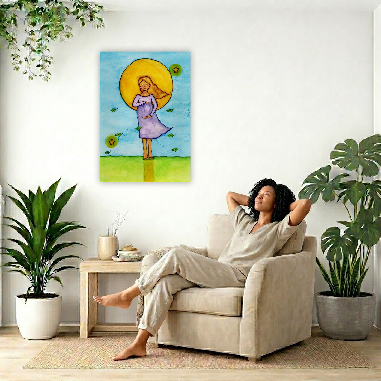 A woman standing in meditation under a bright yellow sun with a blue sky background, canvas watercolor print. The canvas is in the center of the living room wall. To the right a woman is relaxing in a chair with her hands behind her head. To the left is a green floor plant and a hanging green plant. To the right of her is a green floor plant.
