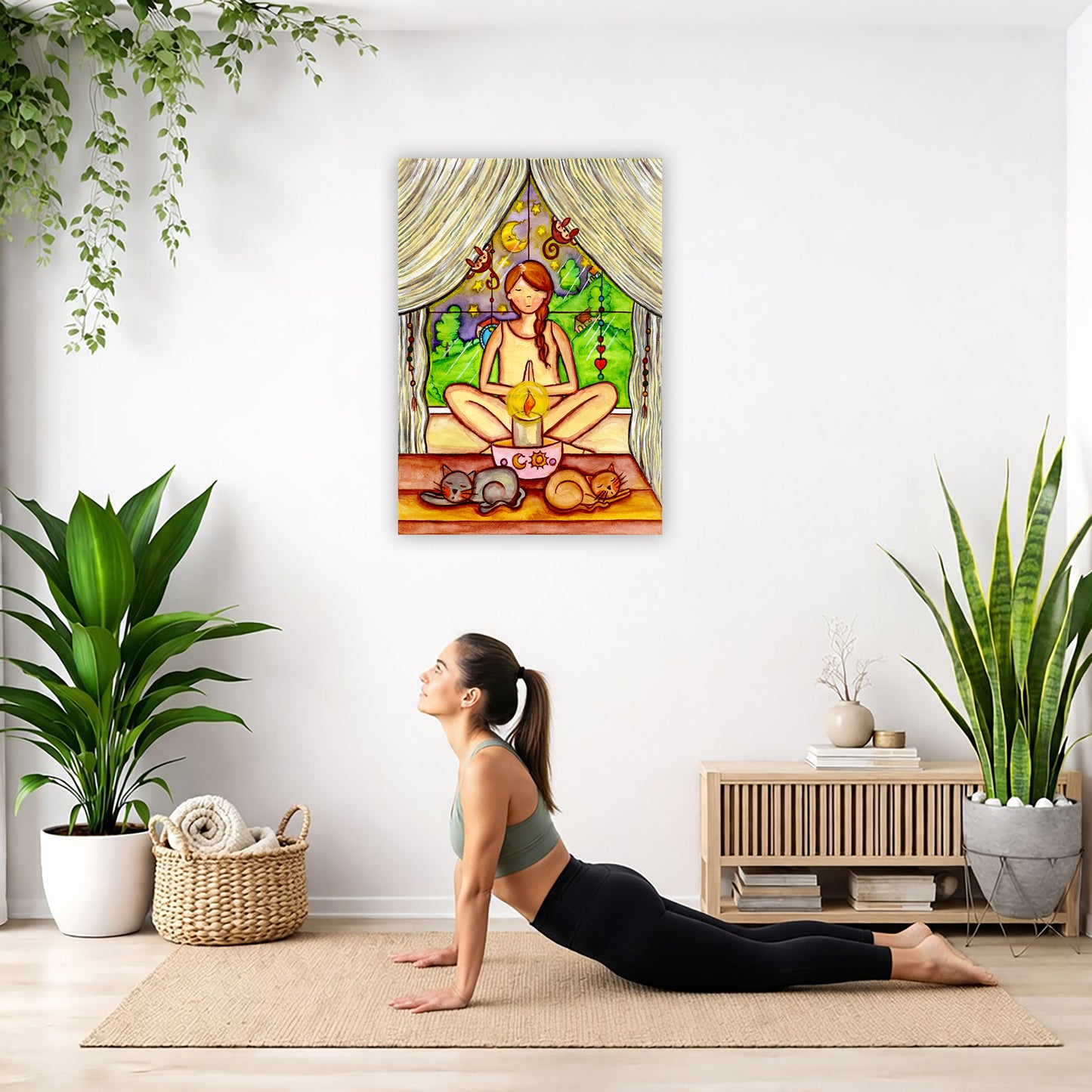 A woman candlelight trataka meditation design on mindfulness canvas.  The canvas is hanging on the wall. A woman on her yoga mat is doing the cobra pose in front of the poster. There is a potted plant, a hanging plant and a basket of cloth material on the left side. On the right side of her is a small cabinet books and a dry plant. There is also a floor plant on the right side.
