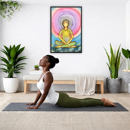 Framed poster of a man's heart chakra glowing and the colorful aura energy surrounding him.  The poster is hanging on the wall. A woman on her yoga mat is doing the cobra pose in front of the poster. There is a potted plant and a hanging plant on the left side. A padded bench with material draped over it is behind her. On the right side are two potted plants.
