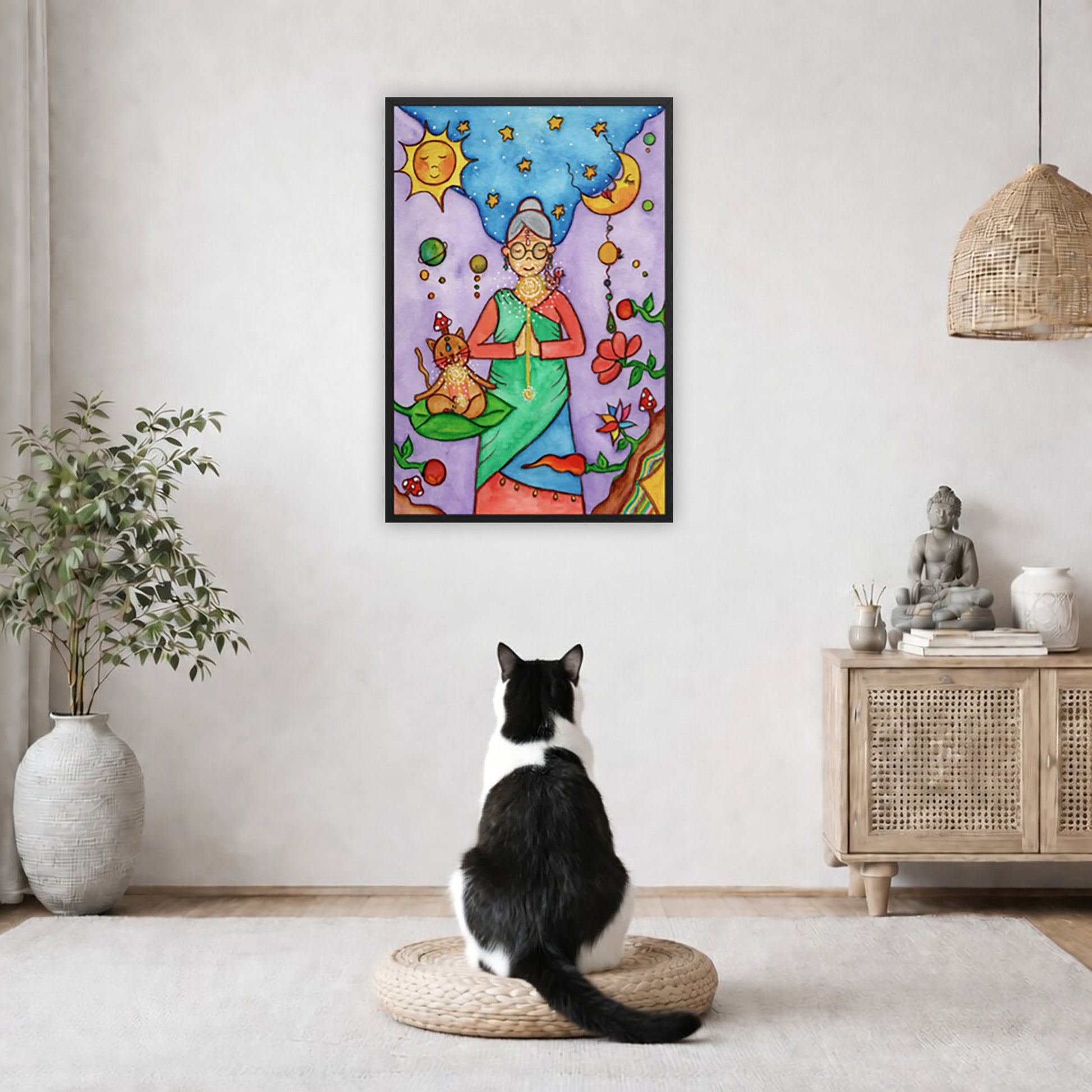 Framed poster of woman in standing prayer meditation with glowing throat chakra under sun and moon. Her cat is meditating beside her.  The poster is in the center of the living room wall. A black and white cat is staring at the poster. There is a potted plant on the left side and a cabinet with a Buddha statue on it on the right side. 
