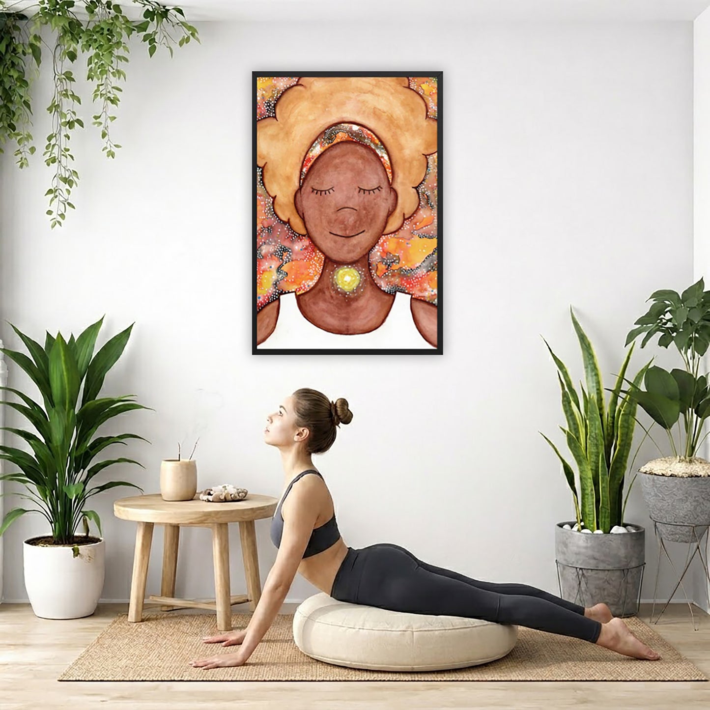 Framed poster of a woman mediating with a glowing throat chakra.  The poster is hanging on the wall. A woman on her yoga pillow is doing the cobra pose in front of the poster. There are several potted plants behind her including a hanging plant on the left side. There is a small table to the woman's left with incense and a bowl of shells on it.