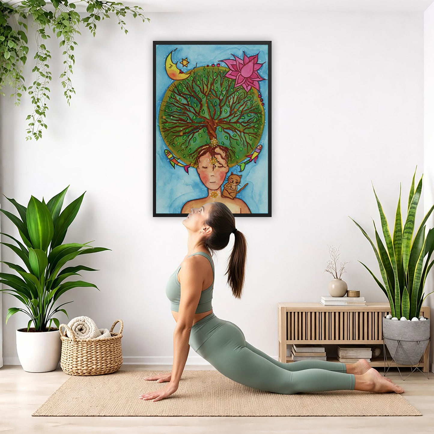 Framed poster of the crown chakra of a woman opening up like the tree of life. A cat is on her shoulder.  The poster is hanging on the wall. A woman on her yoga mat is doing the cobra pose in front of the poster. There is a potted plant and a hanging plant on the left side. A padded bench with material draped over it is behind her. On the right side are two potted plants.
