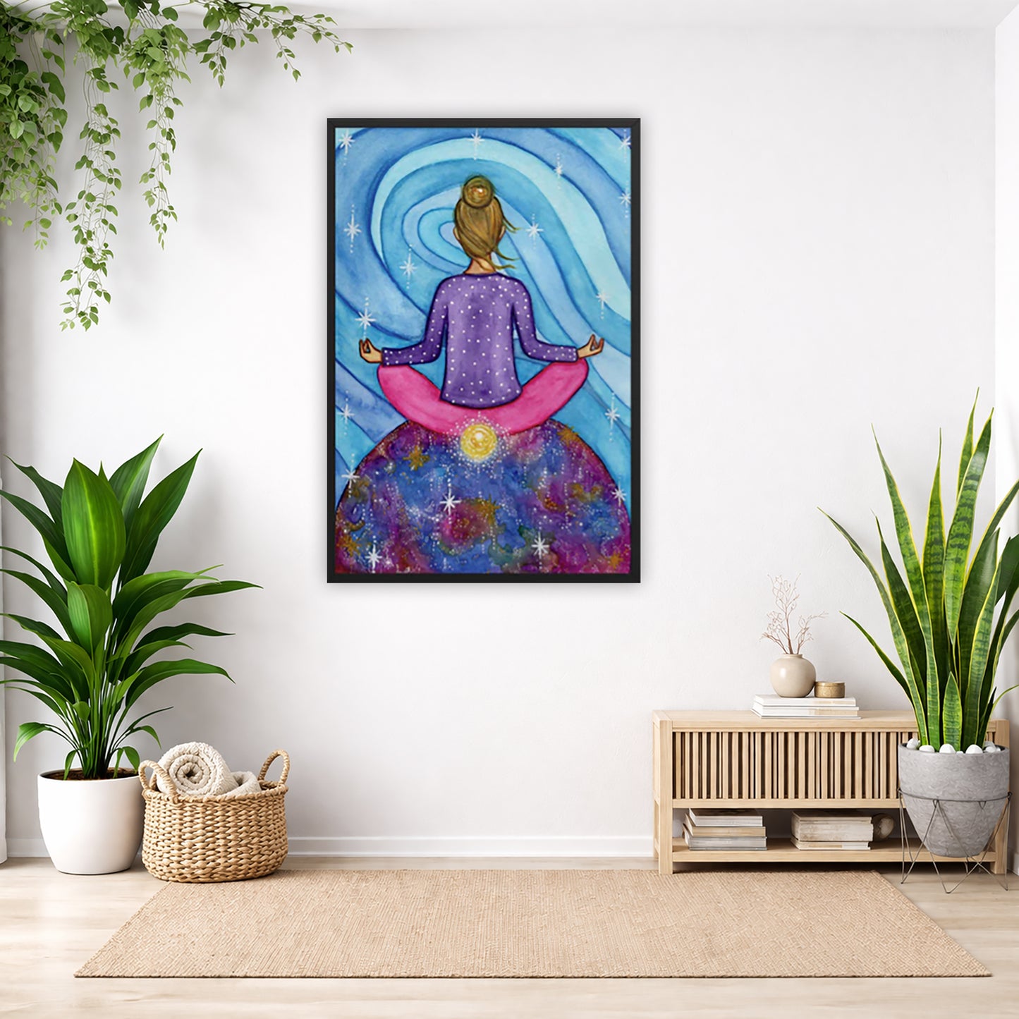 Framed poster of a woman meditating on top of the world with her root chakra glowing.  The poster is in the center of the living room wall. Below the poster to the left is a basket of material and a green floor plant. There is also a green hanging plant. To the right is a small table with books, a candle and a vase with dry flora. There is also a large green floor plant.
