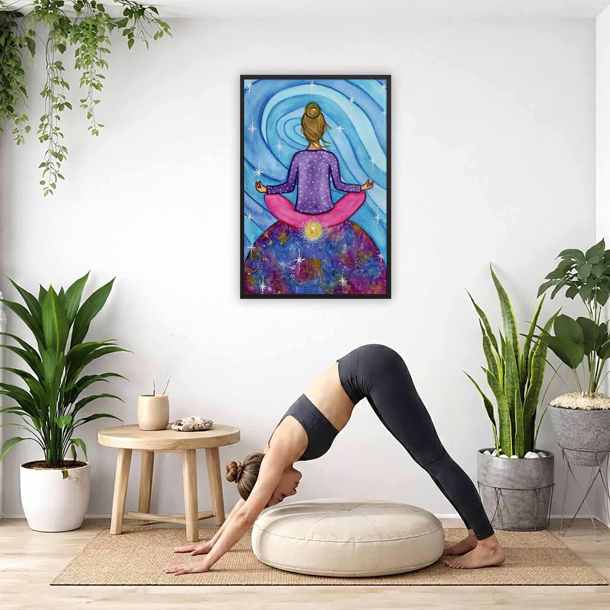 Framed poster of a woman meditating on top of the world with her root chakra glowing. The poster is in the center of the living room wall. Below the poster a woman is doing the yoga posture downward facing dog on a yoga mat.