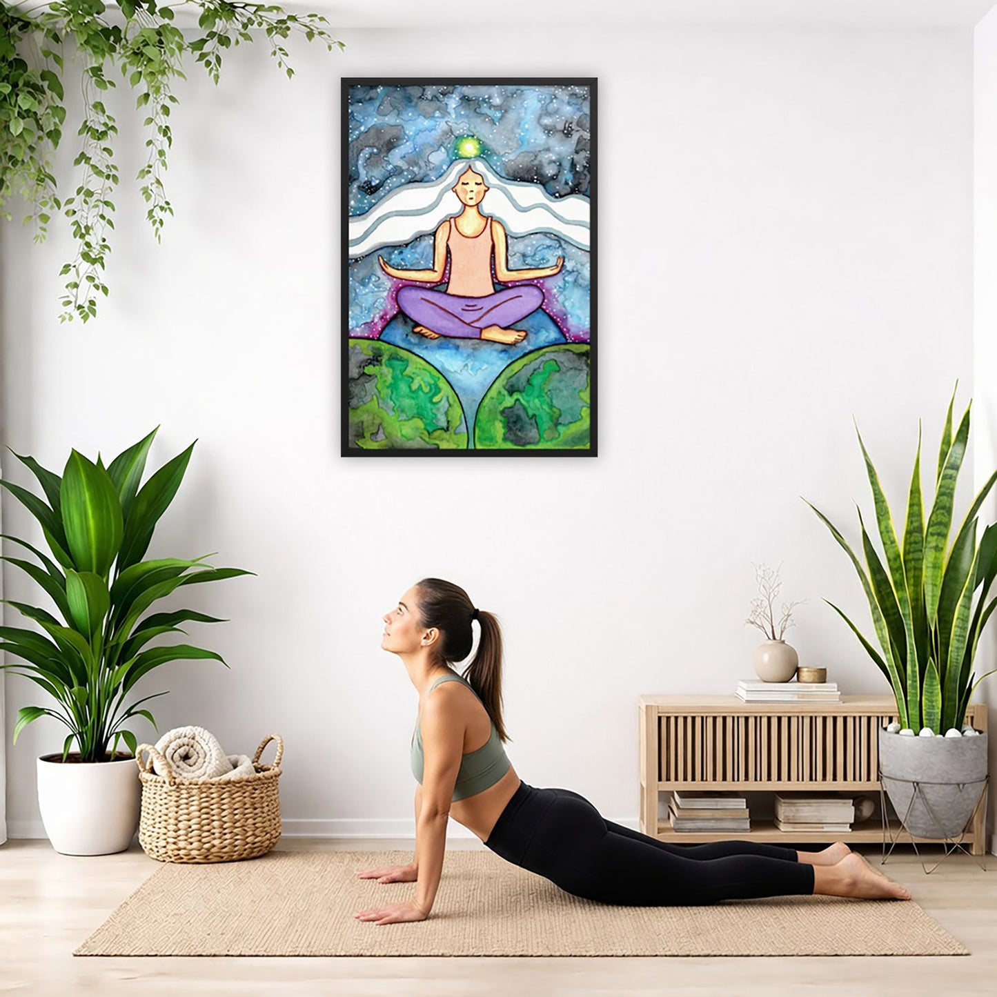 Framed poster of a meditating woman with her crown chakra glowing. The poster is hanging on the wall. A woman on her yoga mat is doing the cobra pose in front of the poster. There is a potted plant and a hanging plant on the left side. A padded bench with material draped over it is behind her. On the right side are two potted plants.
