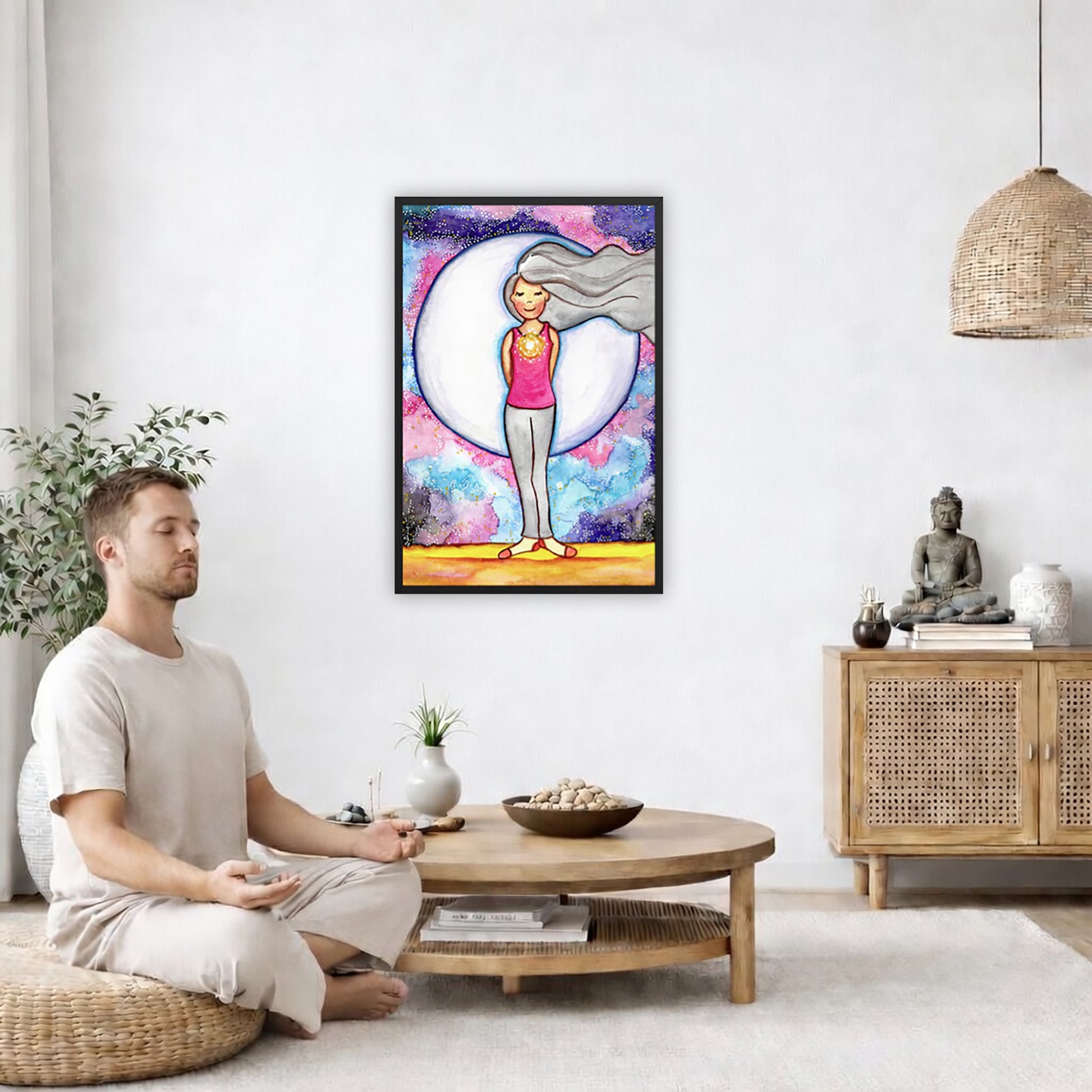 Framed poster of a woman's heart chakra glowing with beautiful love energy. The poster is in the center of the living room wall. A man is on a chair on the left side of his living room meditating. On the right side is a cabinet with a Buddha statue on it.
