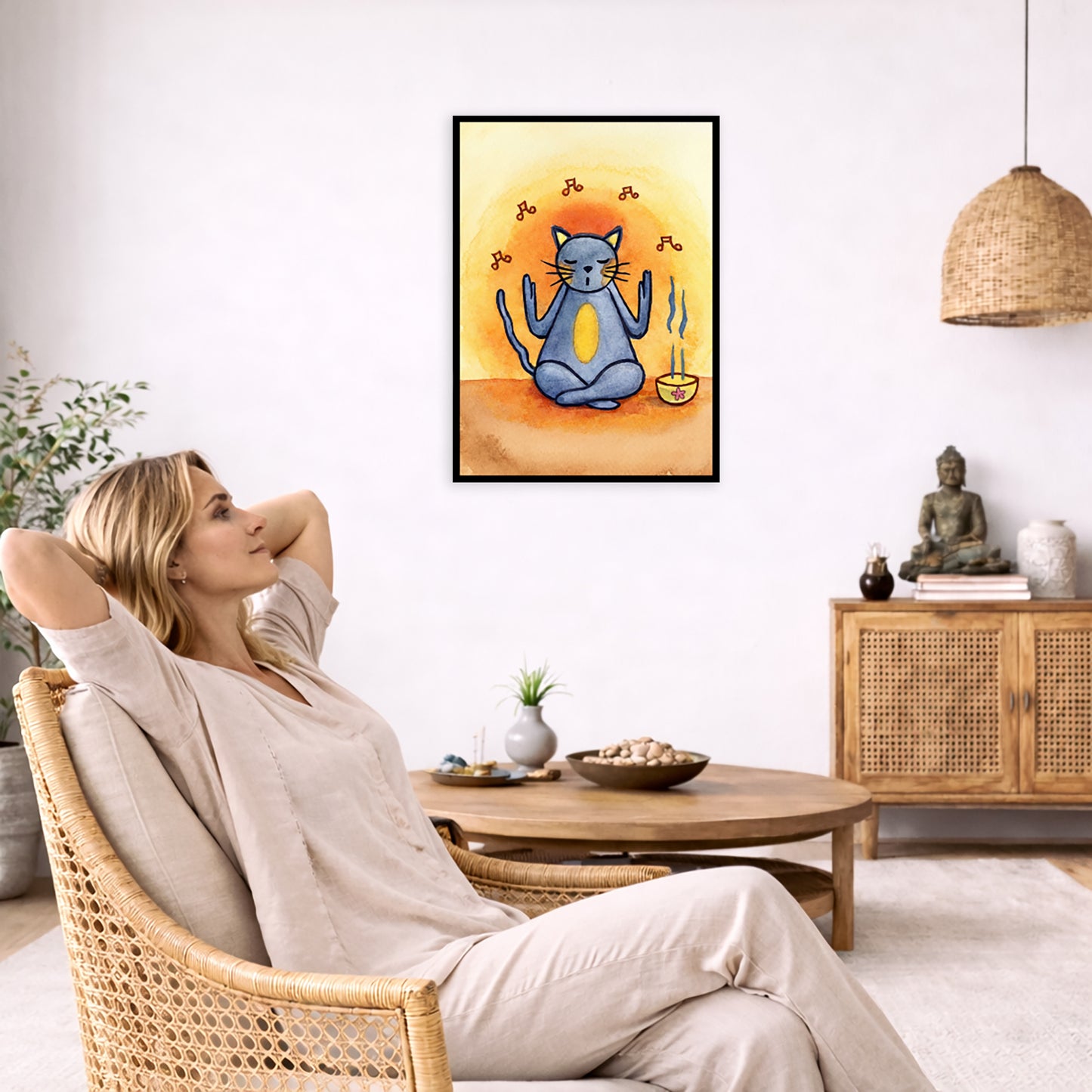 Gray cat chants with incense burning framed poster.  This is a print of the original hand-painted watercolor by Brazilian artist Thaís Suizo.  The poster is in the center of the living room wall. To the left a woman is relaxing in her chair with her hands behind her head. There is a small coffee table beside her with a bowl of walnuts, incense and a small green plant. On the right side of the room is a cabinet with a Buddha statue, books and an urn on it. To the left is a green floor plant.
