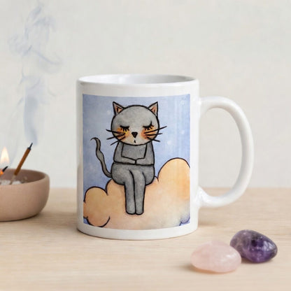Gray cat Meditating on an orange cloud mindfulness coffee mug. The mug is on a wicker mat. Beside the mug are meditation stones. Behind the mug incense is burning.