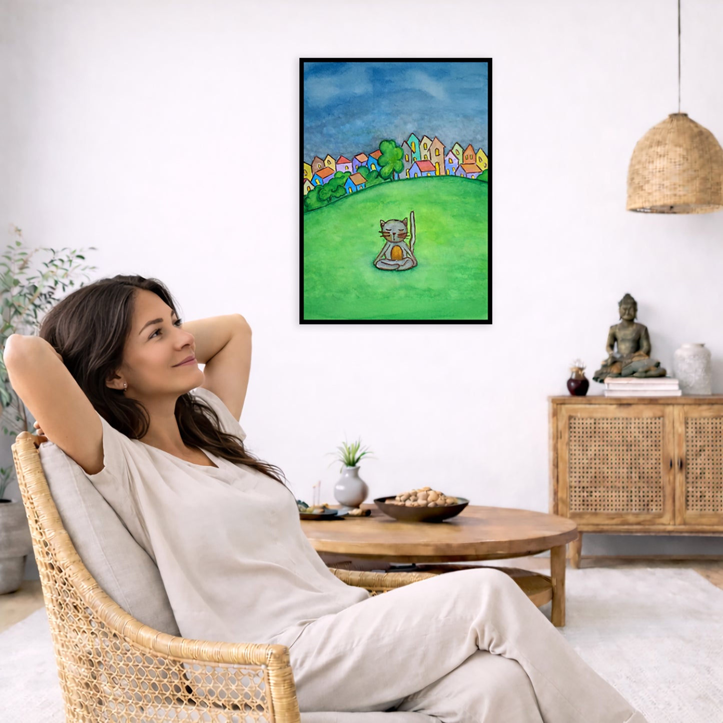 A gray cat meditating in the park framed poster. This is a print of the original hand-painted watercolor by Brazilian artist Thaís Suizo.  The poster is in the center of the living room wall. To the left a woman is relaxing in her chair with her hands behind her head. There is a small coffee table beside her with a bowl of walnuts, incense and a small green plant. On the right side of the room is a cabinet with a Buddha statue, books and an urn on it. To the left is a green floor plant.