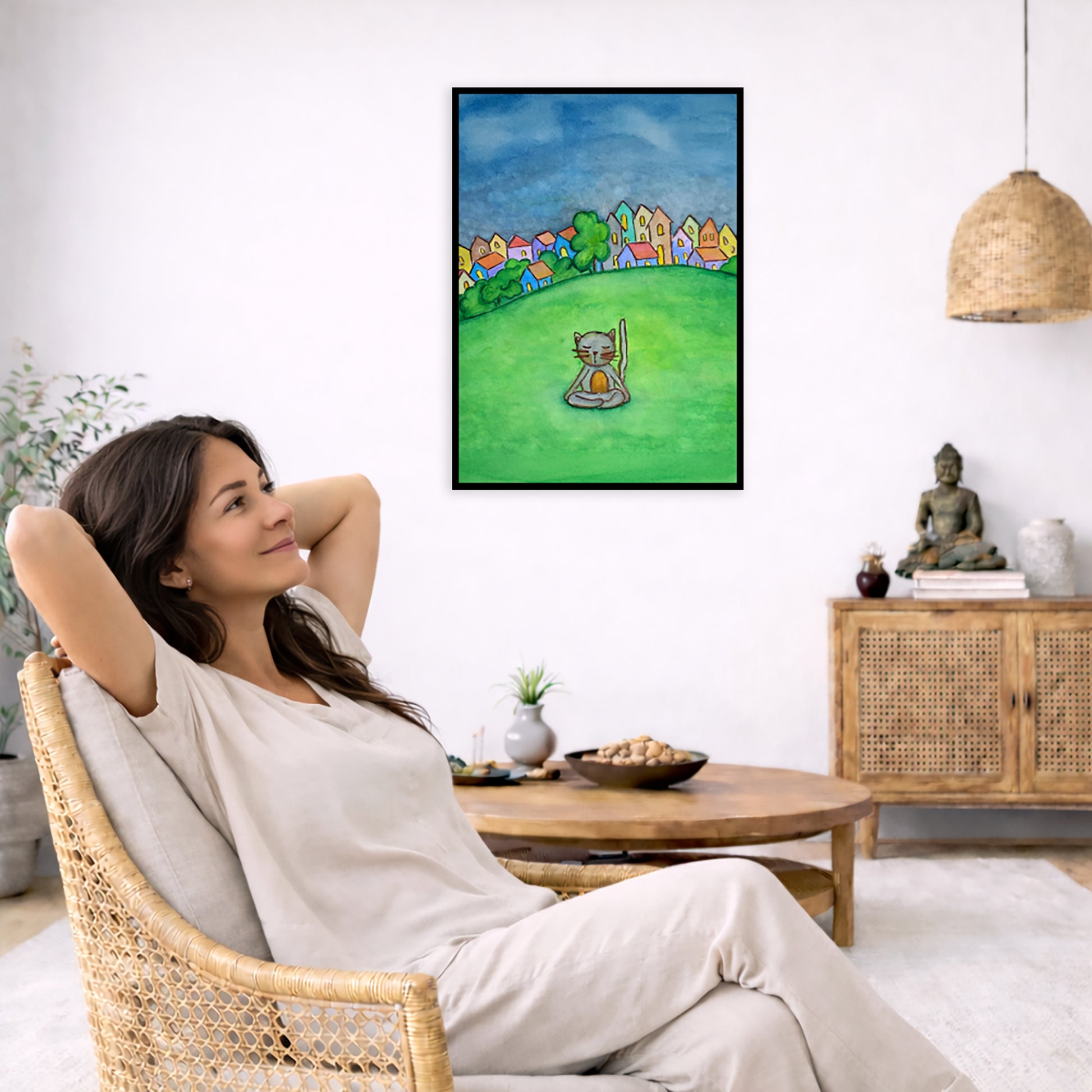 A gray cat meditating in the park framed poster. This is a print of the original hand-painted watercolor by Brazilian artist Thaís Suizo.  The poster is in the center of the living room wall. To the left a woman is relaxing in her chair with her hands behind her head. There is a small coffee table beside her with a bowl of walnuts, incense and a small green plant. On the right side of the room is a cabinet with a Buddha statue, books and an urn on it. To the left is a green floor plant.