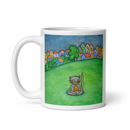 Gray cat meditating peacefully in a park surrounded by a large lawn and a row of houses in the distant background coffee mug. This illustration of a cat meditating is on both sides of the mug. In this image the handle of the mug is on the left side.