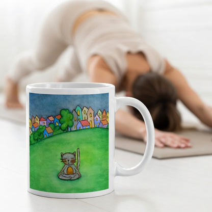 Gray cat meditating peacefully in a park surrounded by a large lawn and a row of houses in the distant background coffee mug.  The mug is in front of a woman doing the yoga posture downward facing dog.