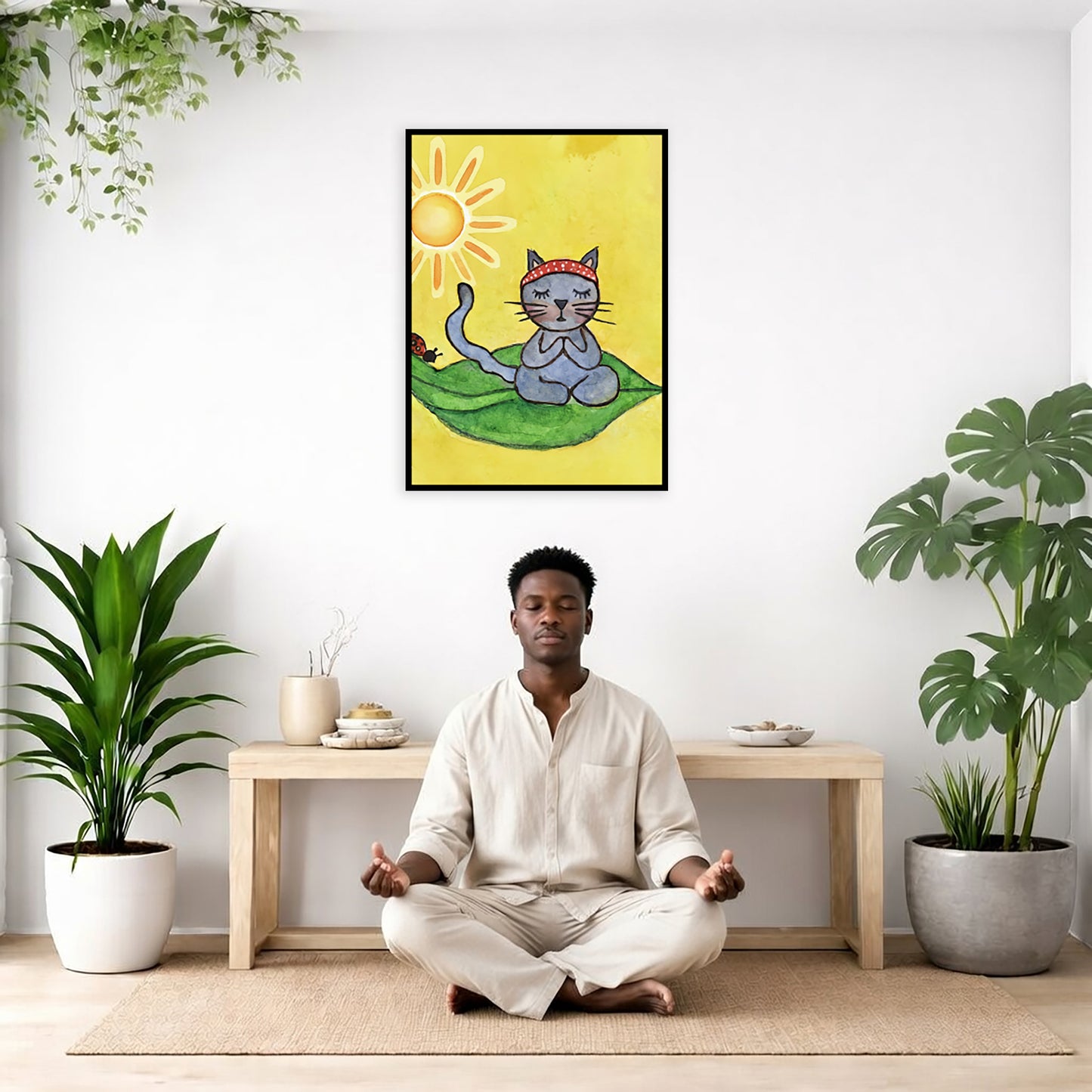 Gray cat meditating on a leaf under a sunny sky framed poster. This is a print of the original hand-painted watercolor by Brazilian artist Thaís Suizo. The poster is framed hanging on a living room wall. A man is mediating facing the poster. There is a coffee table in front of him with burning candles and a Buddha statue. There are green potted plants on each side of the room.
