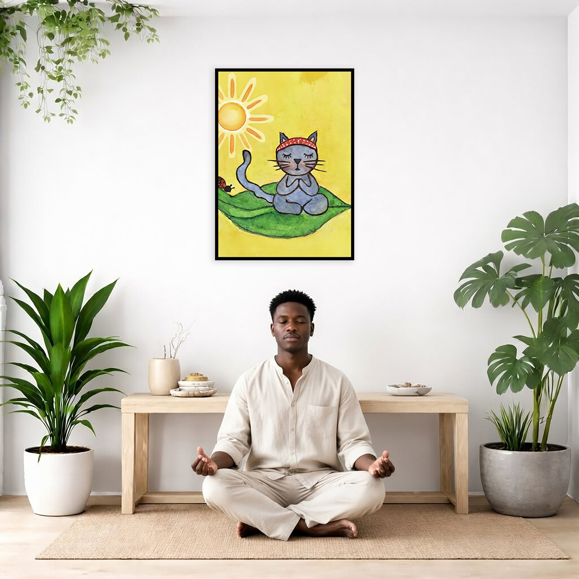 Gray cat meditating on a leaf under a sunny sky framed poster. This is a print of the original hand-painted watercolor by Brazilian artist Thaís Suizo. The poster is framed hanging on a living room wall. A man is mediating facing the poster. There is a coffee table in front of him with burning candles and a Buddha statue. There are green potted plants on each side of the room.
