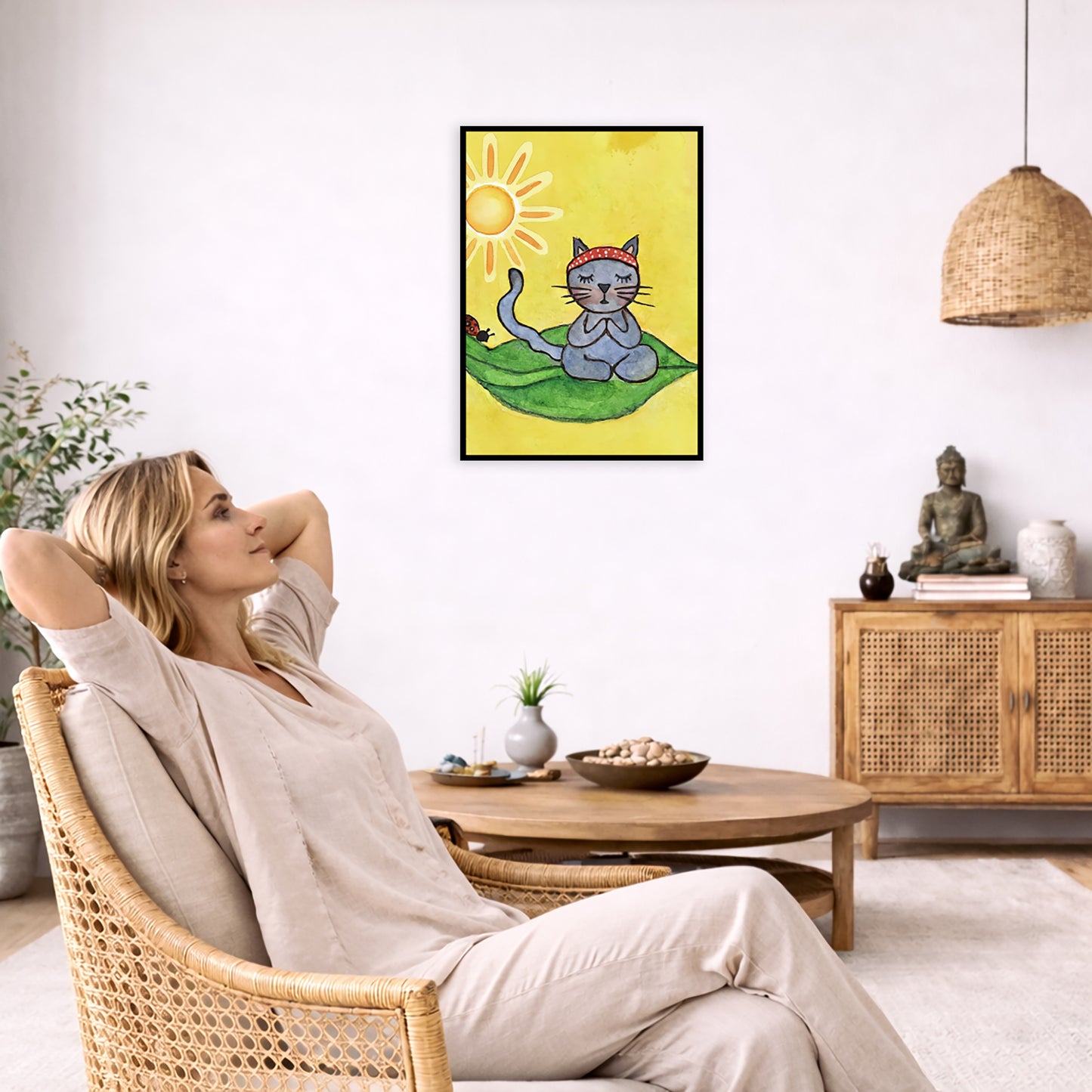 Gray cat meditating on a leaf under a sunny sky framed poster. This is a print of the original hand-painted watercolor by Brazilian artist Thaís Suizo.  The poster is in the center of the living room wall. To the left a woman is relaxing in her chair with her hands behind her head. There is a small coffee table beside her with a bowl of walnuts, incense and a small green plant. On the right side of the room is a cabinet with a Buddha statue, books and an urn on it. To the left is a green floor plant.