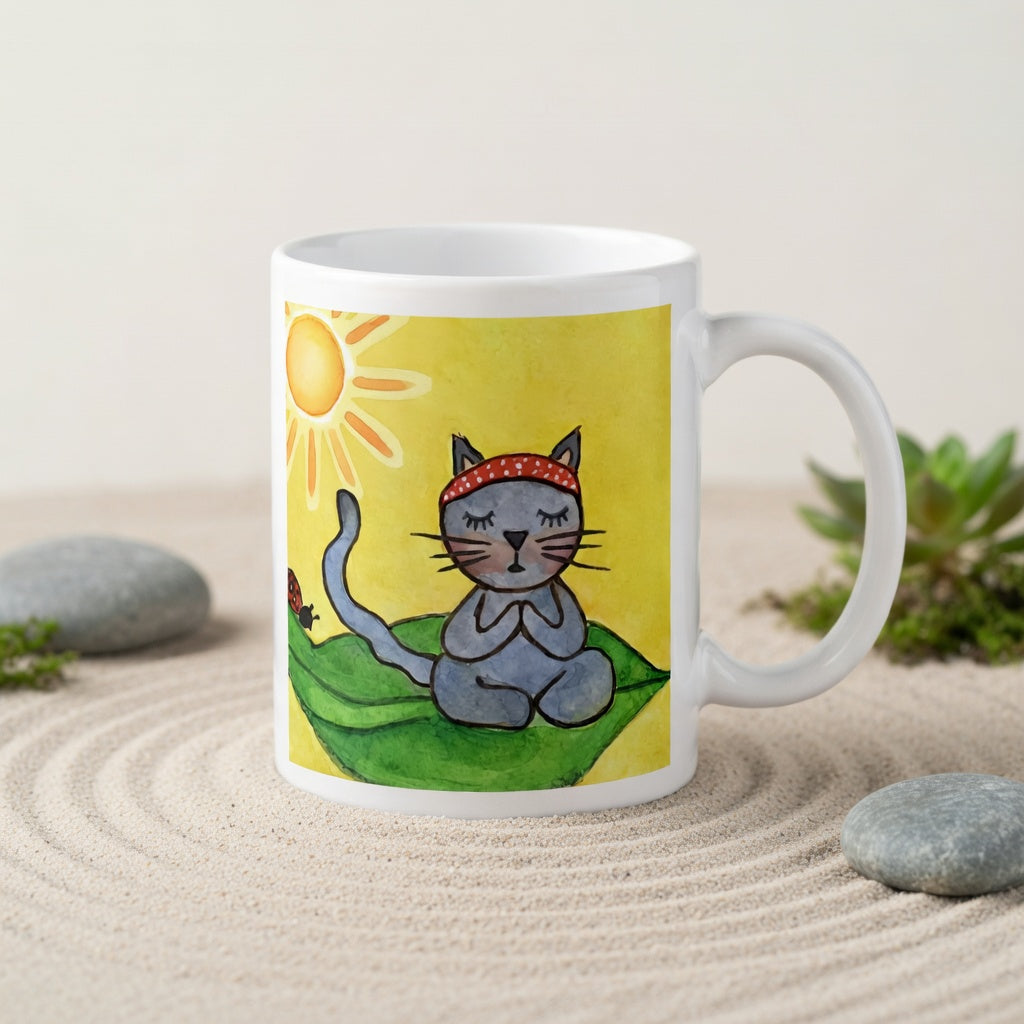 Gray cat meditating peacefully on a leaf in the bright sunlight meditation mug.  The mug is in a beautifully raked zen garden with bushes and plants behind it.