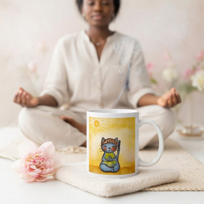 Gray cat meditating on a sunny day mindfulness mug.  The mug is sitting on a white mat with a flowers  beside it. A woman is meditating in the background.