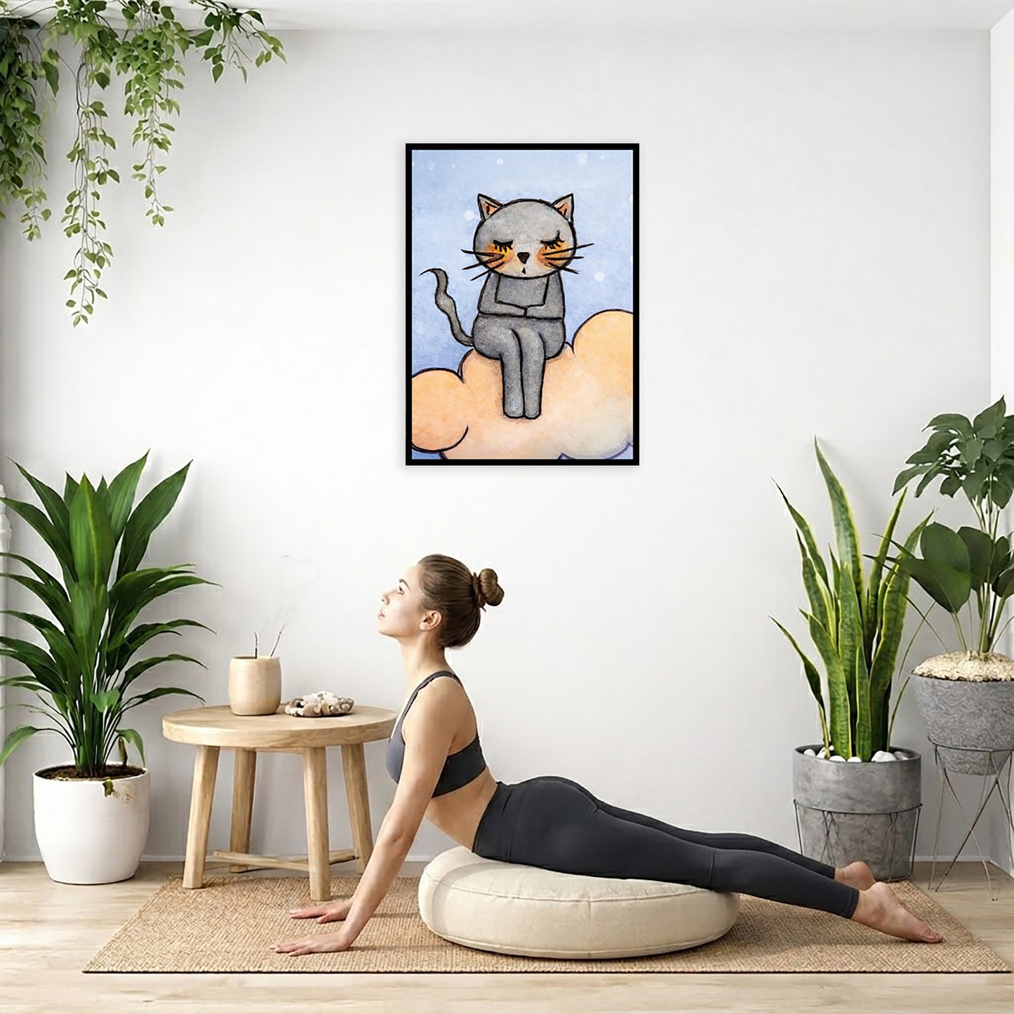 A gray cat meditating on an orange cloud framed poster. This is a print of the original hand-painted watercolor by Brazilian artist Thaís Suizo.  The poster is hanging on the wall. A woman on her yoga pillow is doing the cobra pose in front of the poster. There are several potted plants behind her including a hanging plant on the left side. There is a small table to the woman's left with incense and a bowl of shells on it.