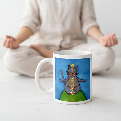 Gray cat meditating with glowing chakras on a deep blue calming background.  The mug is on the floor at the bottom center of the image. In the background a woman is meditating. 