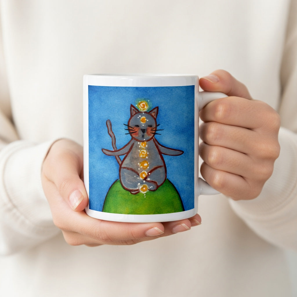 Gray cat meditating with glowing chakras on a deep blue calming background. The hands of a person wearing a white shirt are holding the mug up to display the design.