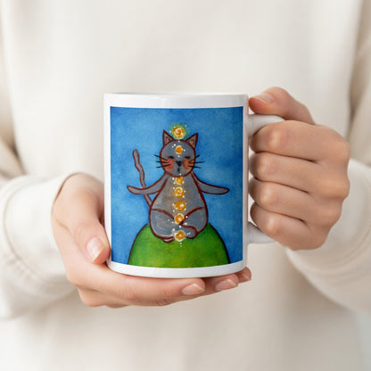 Gray cat meditating with glowing chakras on a deep blue calming background. The hands of a person wearing a white shirt are holding the mug up to display the design.