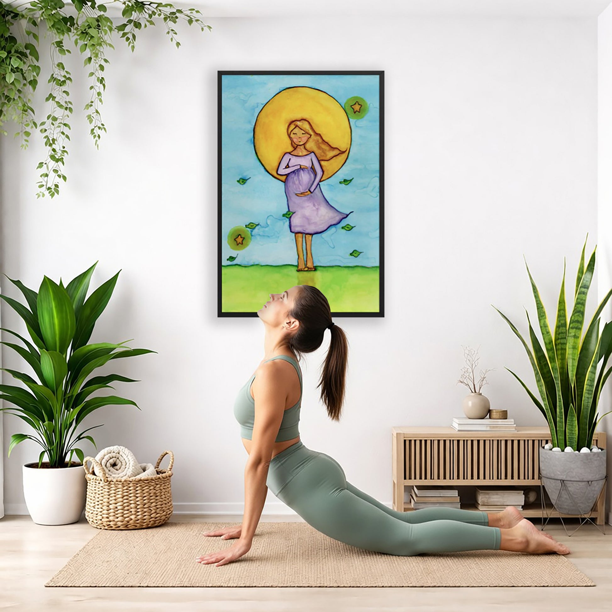 Woman standing in meditation under a bright sun with soft blue sky background, framed watercolor print.  The poster is hanging on the wall. A woman on her yoga mat is doing the cobra pose in front of the poster. There is a potted plant, a hanging plant and a basket of cloth material on the left side. On the right side of her is a small cabinet books and a dry plant. There is also a floor plant on the right side.