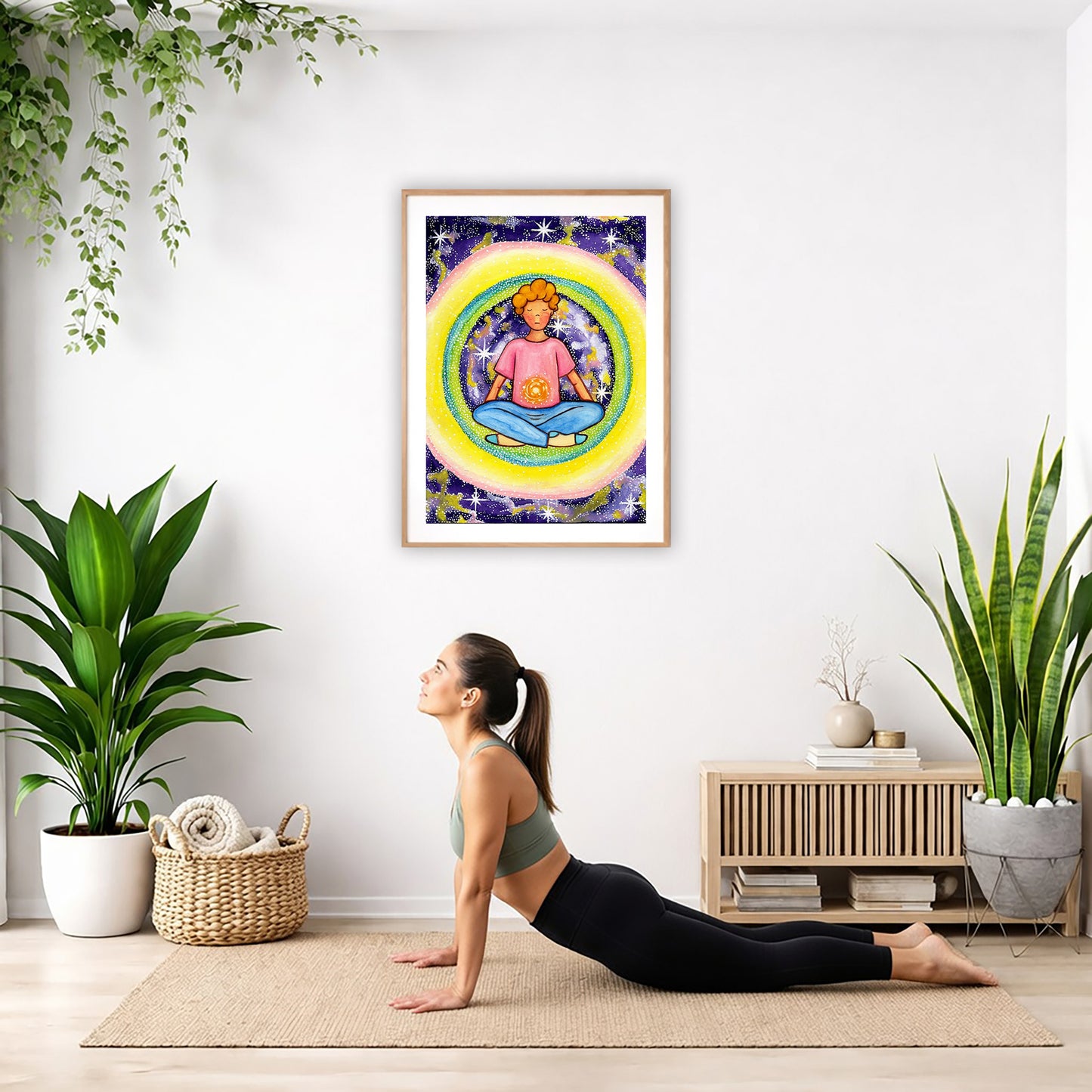 Poster of a young man meditating with his solar plexus glowing. Surrounding him is his colorful aura.  The poster is hanging on the wall. A woman on her yoga mat is doing the cobra pose in front of the poster. There is a potted plant and a hanging plant on the left side. A padded bench with material draped over it is behind her. On the right side are two potted plants.
