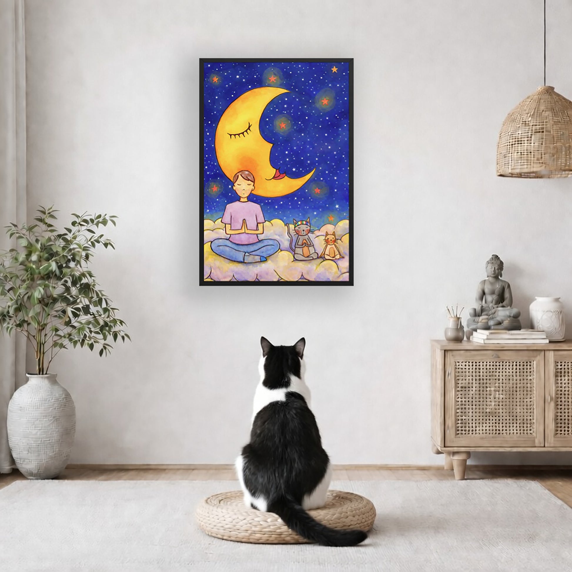 A man and his cats meditate together sitting on a cloud under the moon and the night sky. The poster is framed and is in the center of the living room wall. A black and white cat is staring at the poster. There is a potted plant on the left side and a cabinet with a Buddha statue on it on the right side. 
