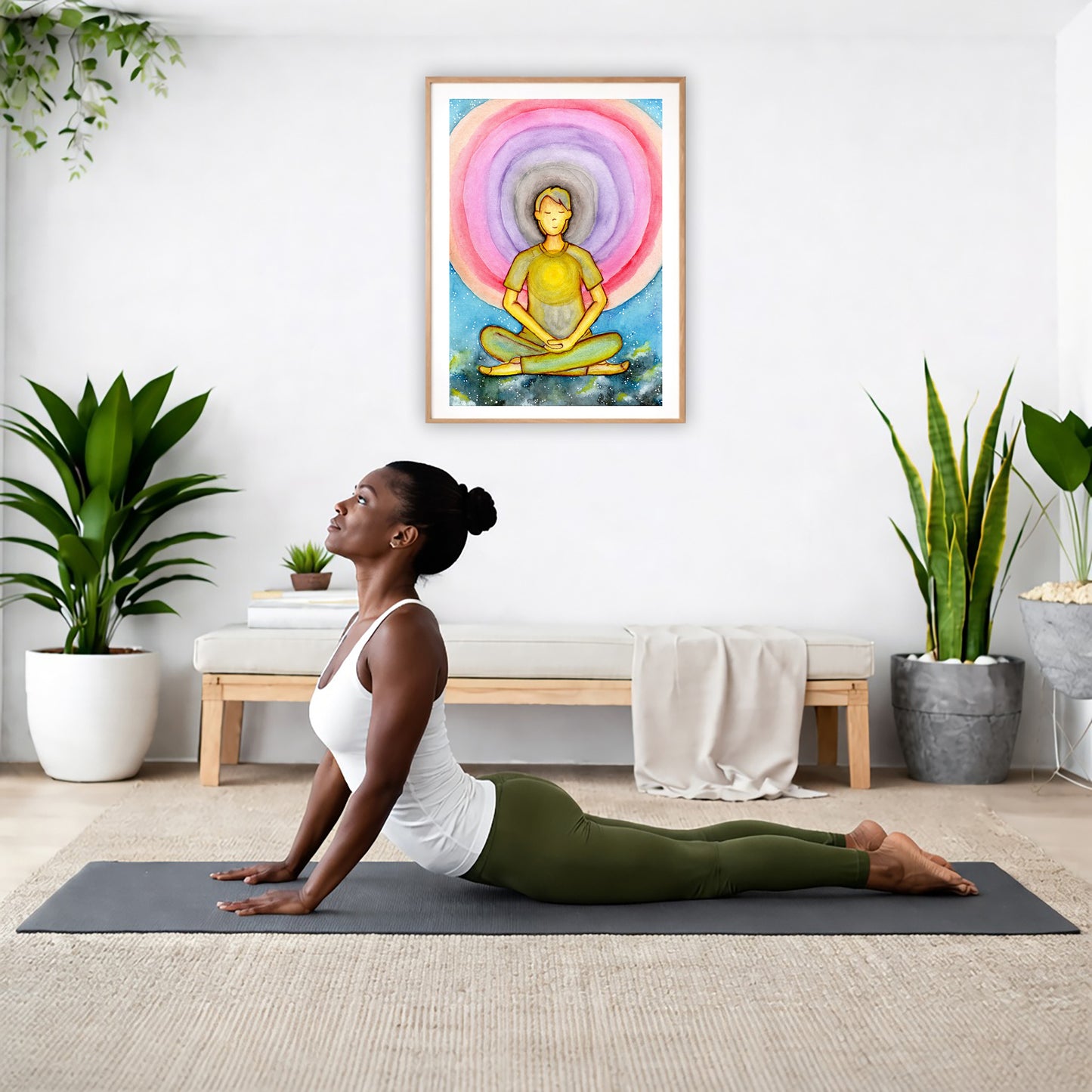 Poster of a young man's heart chakra glowing and aura energy flowing around him.  The poster is hanging on the wall. A woman on her yoga mat is doing the cobra pose in front of the poster. There is a potted plant and a hanging plant on the left side. A padded bench with material draped over it is behind her. On the right side are two potted plants.
