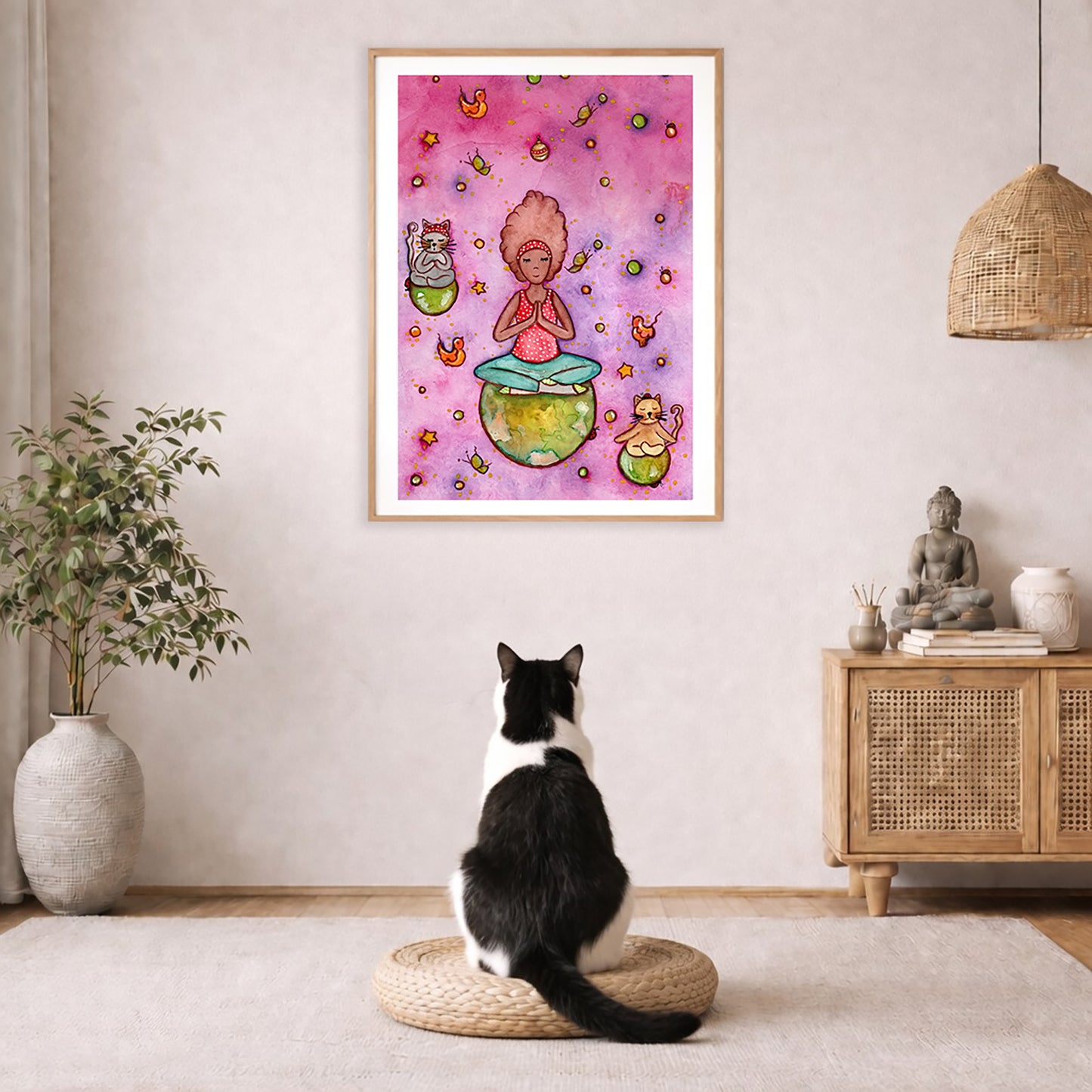 A woman and her cats meditating on the world with a pink background poster. The poster is framed and is in the center of the living room wall. A black and white cat is staring at the poster. There is a potted plant on the left side and a cabinet with a Buddha statue on it on the right side. 
