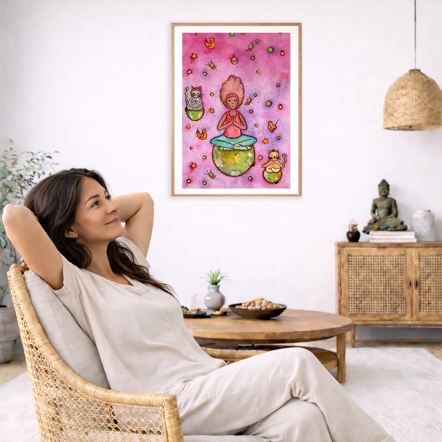 A woman and her cats meditating on the world with a pink background poster.  The poster is framed and is in the center of the living room wall. To the left a woman is relaxing in her chair with her hands behind her head. There is a small coffee table beside her with a bowl of walnuts, incense and a small green plant. On the right side of the room is a cabinet with a Buddha statue, books and an urn on it. To the left is a green floor plant.