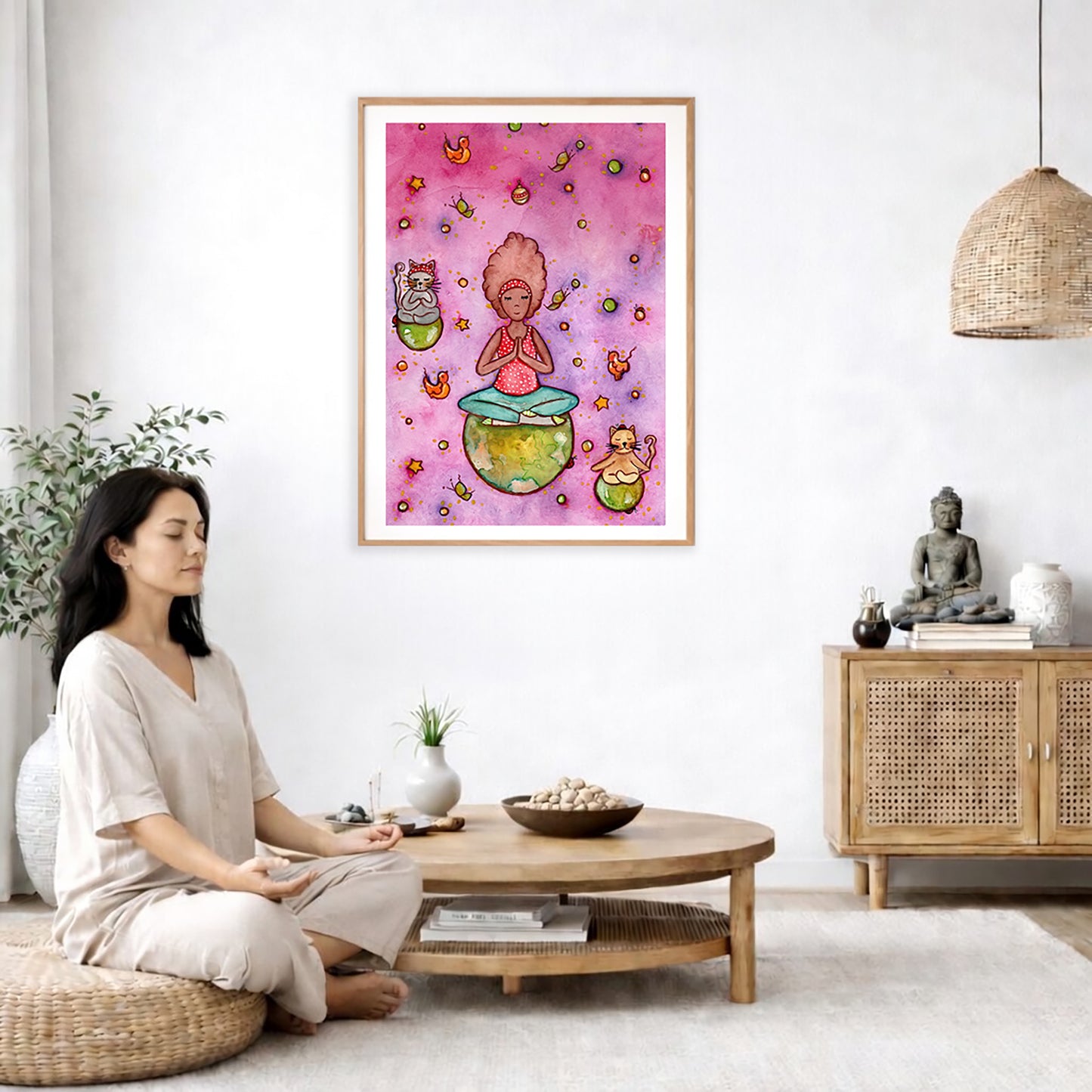 A woman and her cats meditating on the world with a pink background poster.  The poster is framed and is in the center of the living room wall. A woman is on a chair on the left side of his living room meditating. There is a green potted plant behind her. On the right side is a cabinet with a Buddha statue on it.
