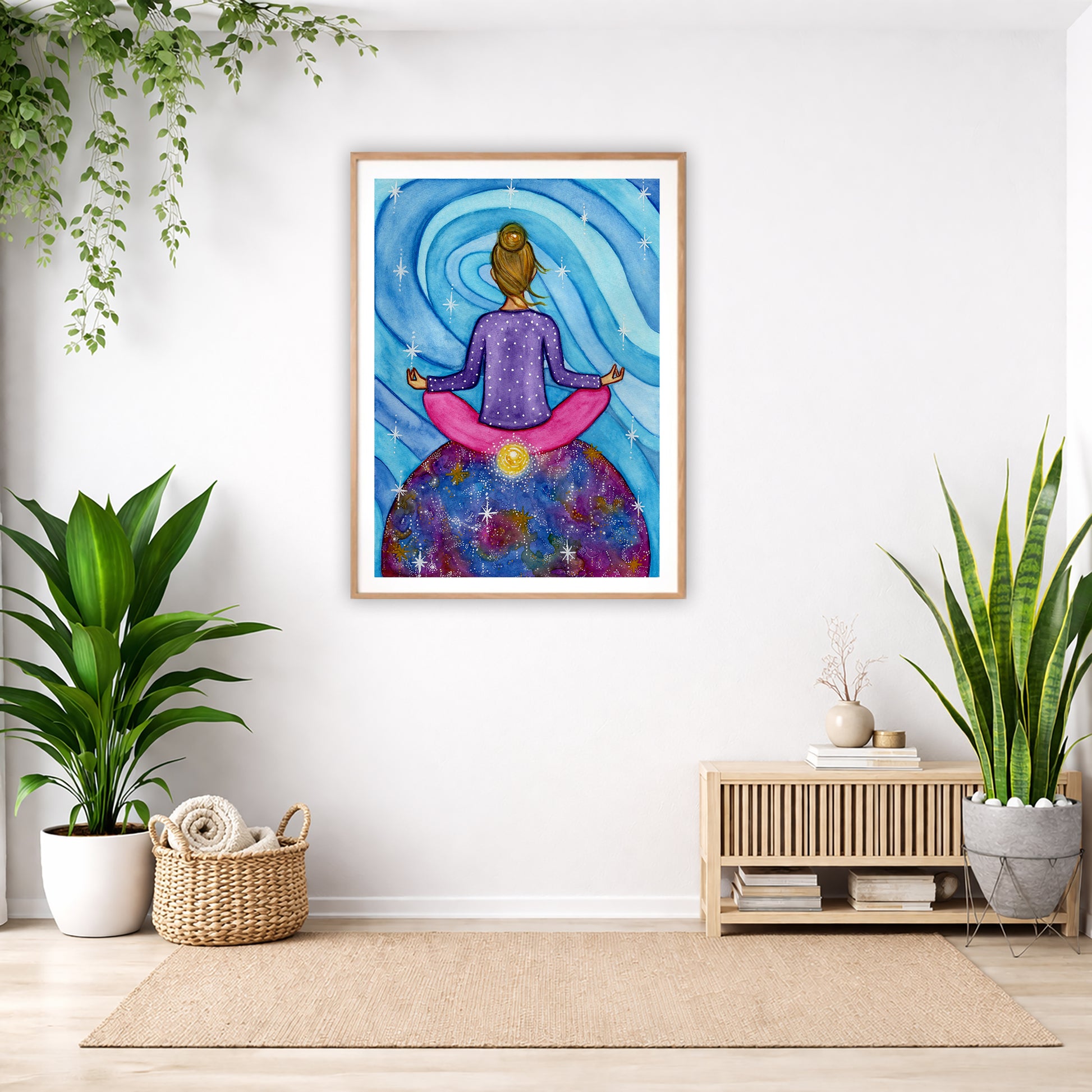 Poster of a woman meditating with her root chakra glowing.  The poster is in the center of the living room wall. Below the poster to the left is a basket of material and a green floor plant. There is also a green hanging plant. To the right is a small table with books, a candle and a vase with dry flora. There is also a large green floor plant.