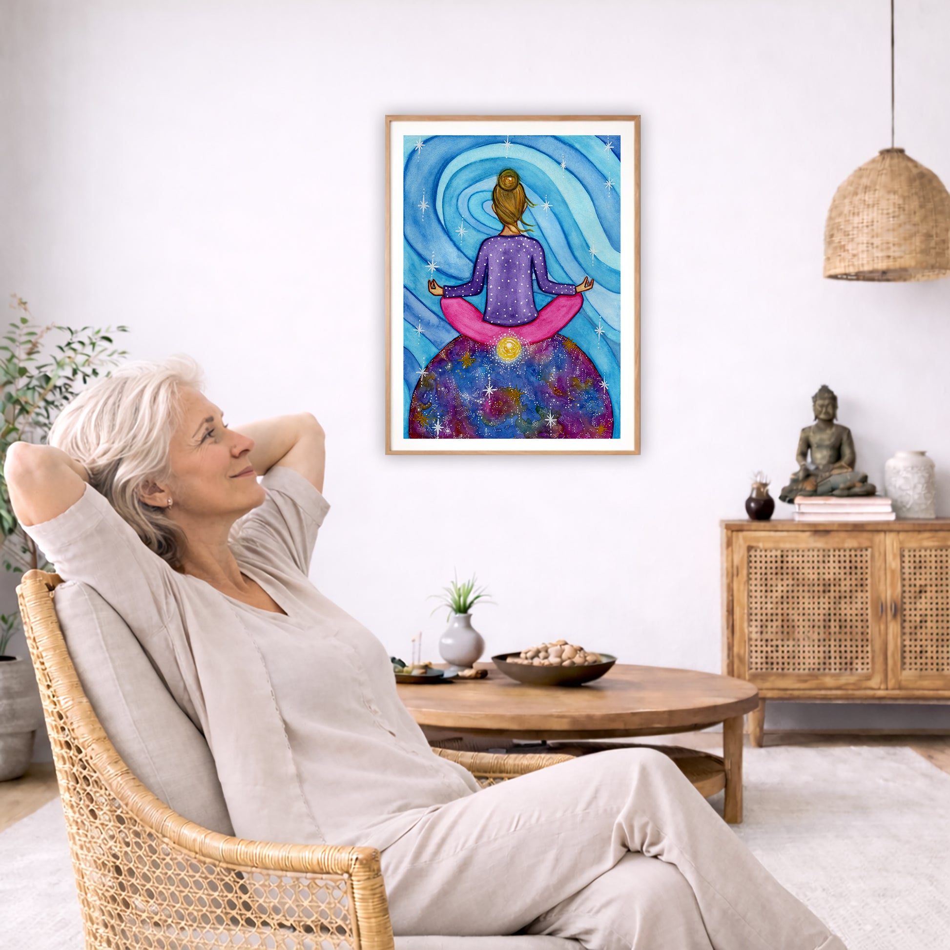 Poster of a woman meditating with her root chakra glowing.  The poster is framed hanging on a living room wall. A woman is mediating facing the poster. There is a coffee table in front of her with burning candles and a Buddha statue. There are green potted plants on each side of the room.
