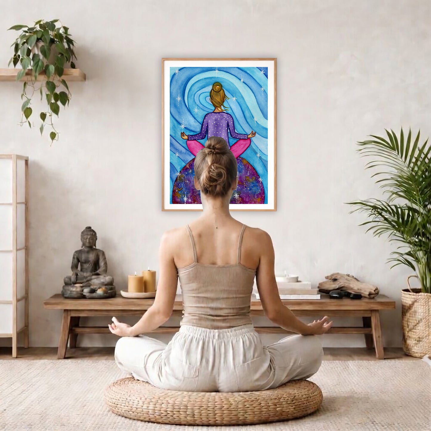 Poster of a woman meditating with her root chakra glowing.  The poster is framed hanging on a living room wall. A woman is mediating facing the poster. There is a coffee table in front of her with burning candles and a Buddha statue. There are green potted plants on each side of the room.
