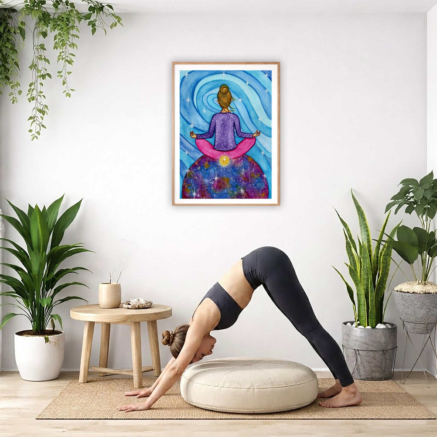 Poster of a woman meditating with her root chakra glowing.  The poster is in the center of the living room wall. Below the poster a woman is doing the yoga posture downward facing dog on a wicker mat.