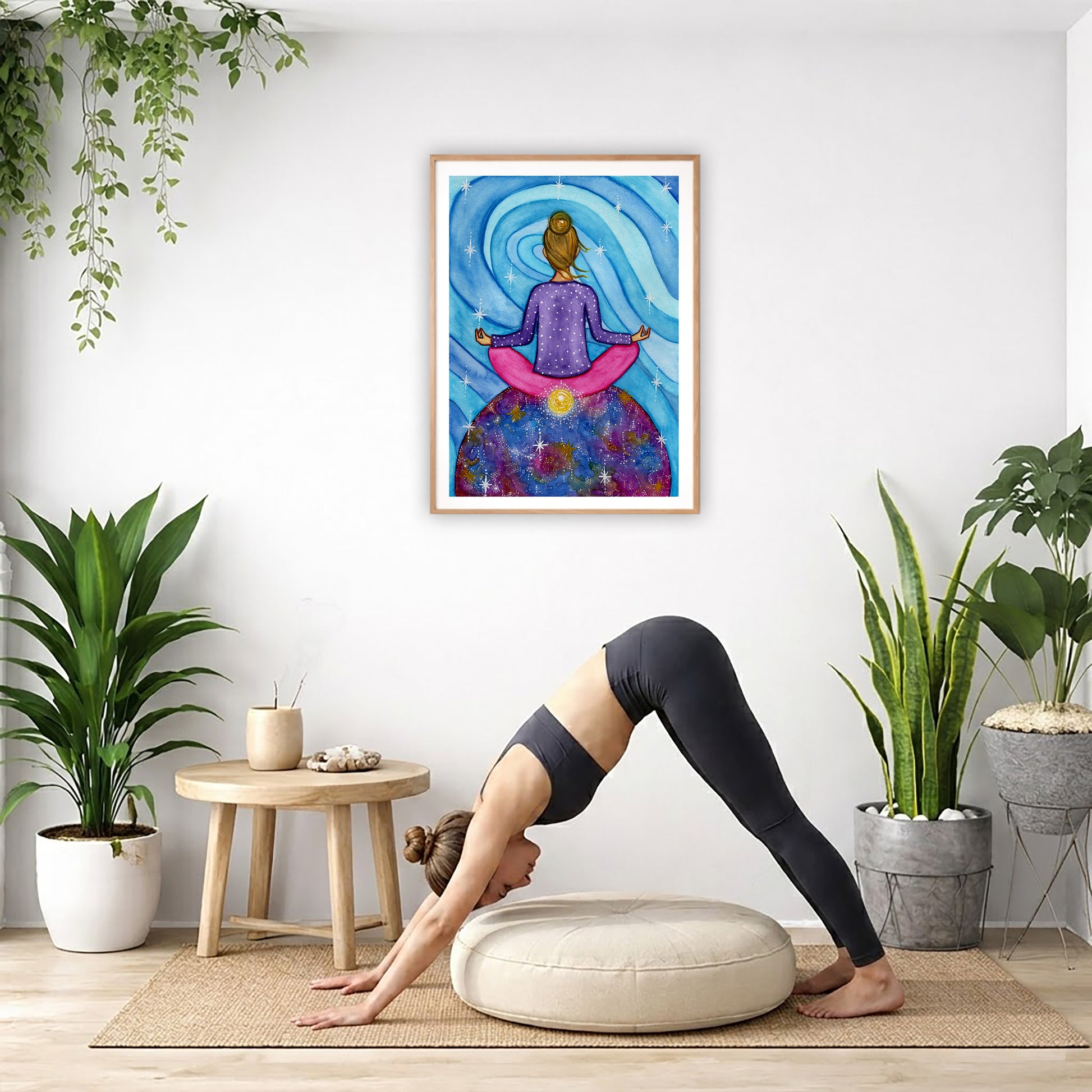Poster of a woman meditating with her root chakra glowing.  The poster is in the center of the living room wall. Below the poster a woman is doing the yoga posture downward facing dog on a wicker mat.