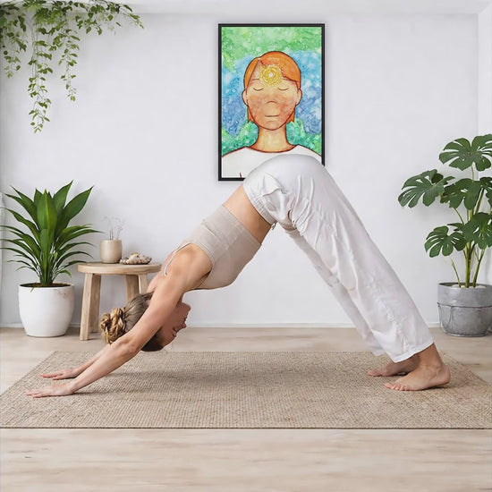 Framed poster of a man with a glowing brow chakra. The poster is in the center of the living room wall. A woman is doing a sun salutation on her yoga mat. There is a coffee table below the poster with burning candles, incense, a Buddha statue and a big piece of driftwood.
