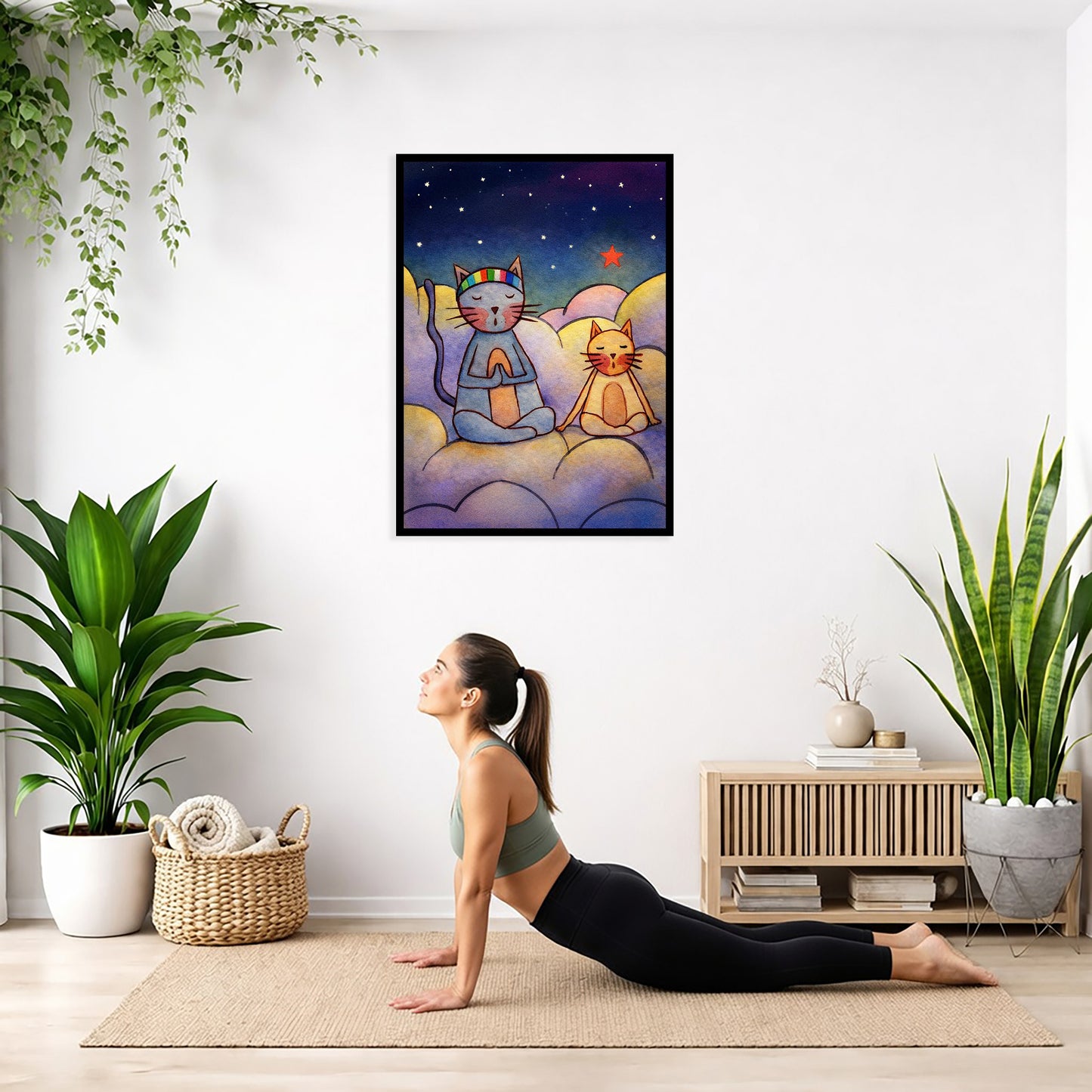 Two cats meditating on a cloud at night under the stars framed poster. This is a print of the original illustration by Brazilian artist Thaís Suizo.  The poster is hanging on the wall. A woman on her yoga mat is doing the cobra pose in front of the poster. There is a potted plant and a hanging plant on the left side. A padded bench with material draped over it is behind her. On the right side are two potted plants.
