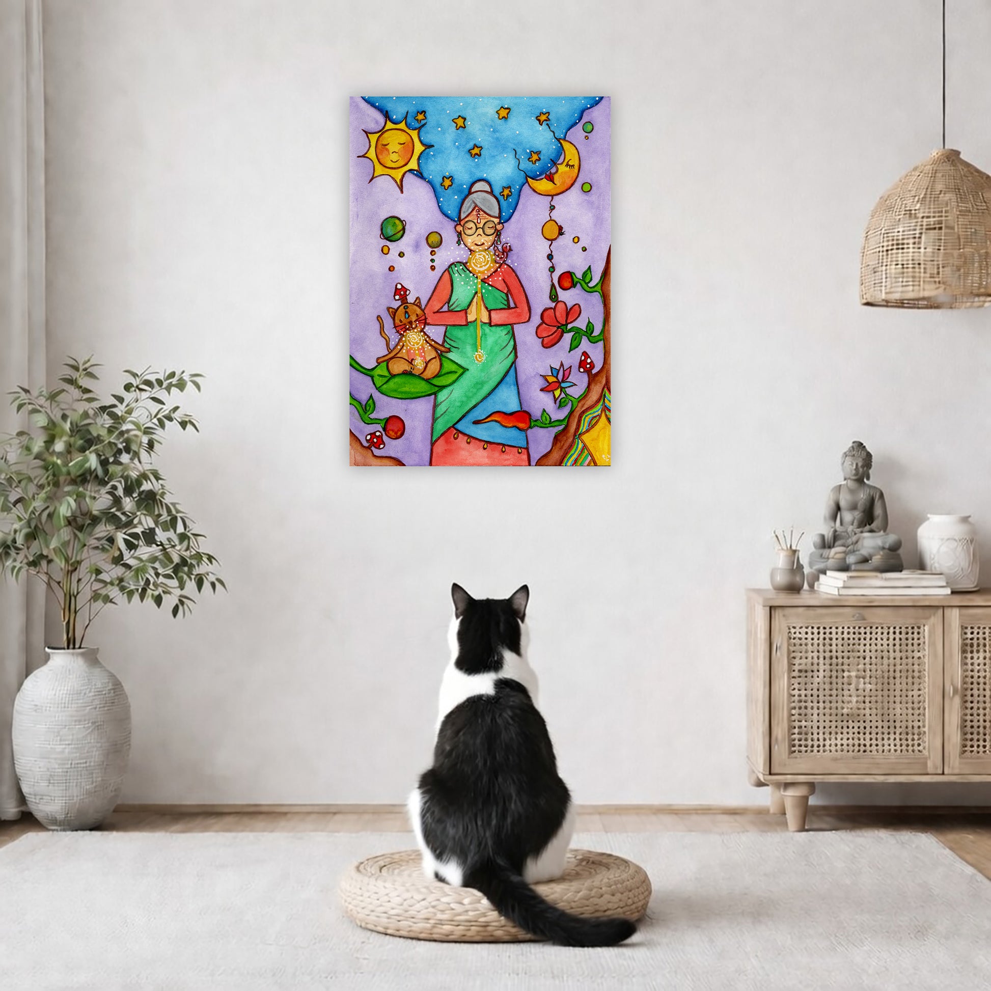 Throat chakra canvas print of a woman and her cat meditating under the sun and the moon.  The poster is in the center of the living room wall. A black and white cat is staring at the poster. There is a potted plant on the left side and a cabinet with a Buddha statue on it on the right side. 
