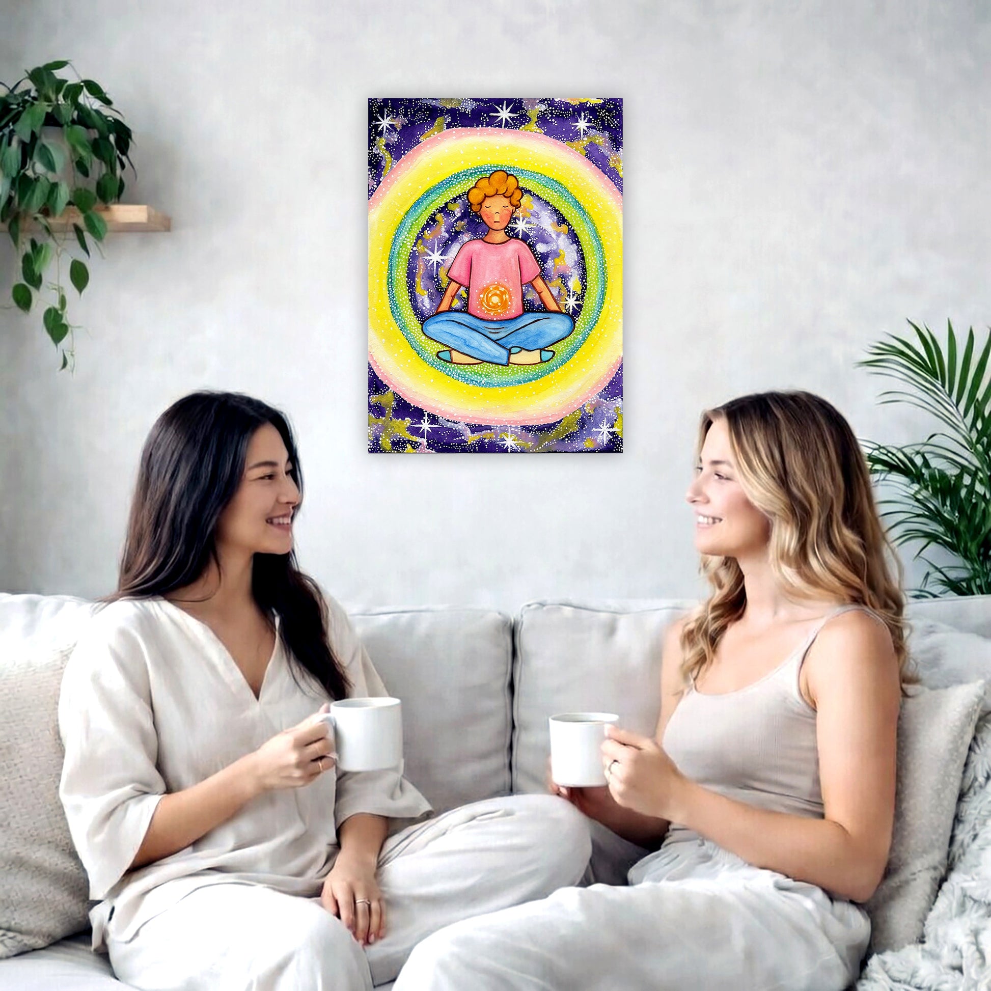 Young man meditating with his solar plexus glowing canvas. Surrounding him is his colorful aura. This is a print of the original hand-painted watercolor by artist Thaís Suizo for the LET'S MEDITATE SERIES of mediation guides. The poster is in the center of the living room wall. Two female friends are having tea, grapes and cookies on a coffee table in front of the poster. On the left there is a green potted plant and to the right there is a hanging lamp.