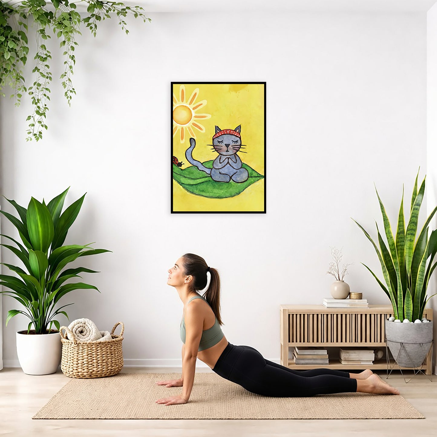 Gray cat meditating on a leaf under a sunny sky framed poster. This is a print of the original hand-painted watercolor by Brazilian artist Thaís Suizo.  The poster is hanging on the wall. A woman on her yoga mat is doing the cobra pose in front of the poster. There is a potted plant on the left side and a cabinet with a Buddha statue on it on the right side.