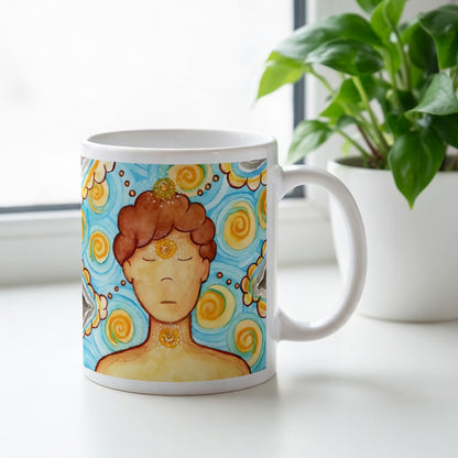 Man meditating with glowing throat, brow and crown chakras, symbolizing communication, intuition and spiritual connection, chakra artwork on a coffee mug. There is a window and a green potted plant behind the mug.
