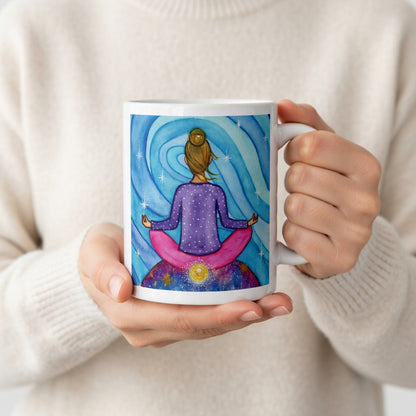 Meditating figure with glowing root chakra at base of spine, symbolizing grounding, stability and security, chakra energy artwork on a coffee mug. The hands of a person wearing a white sweater are holding the mug up to display the design.